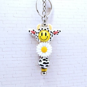 May include: A white and black cow print keyring with a yellow smiley face, a white daisy, and a white and black cow print bead. The keyring is attached to a silver key ring.