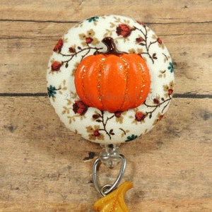 May include: A white fabric button with a floral pattern and a glittery orange pumpkin on top. A gold leaf charm is attached to a clear retractable badge holder.
