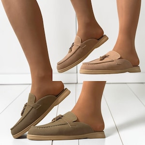 May include: Tan suede slip-on mules with a tassel detail. The shoes have a rounded toe and a low, flat sole. The image shows multiple pairs of the shoes, showcasing their design and style.