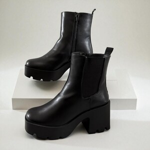 May include: A pair of black platform boots. One boot features a zip closure, the other has elastic side panels. Both boots have a chunky platform sole and a block heel. The boots are made of a smooth, shiny material.