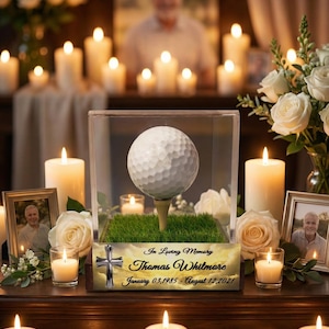 May include: A memorial display featuring a golf ball on a tee inside a clear case, set on a bed of artificial grass. The display includes a sign that reads "In Loving Memory Thomas Whitmore January 03, 1985 - August 12, 2021". Surrounding the display are lit candles and framed photos.