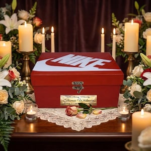 Może przedstawiać: Czerwone prostokątne drewniane pudełko z białym logo Nike na wieczku i złotym metalowym zatrzaskiem. Złota tabliczka z przodu zawiera napis In Loving Memory Thomas Whitmore January 01, 1985 - August 12, 2021, umieszczone na białej koronkowej serwecie.