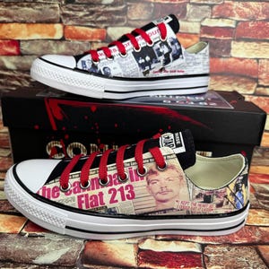 以下が含まれることがあります： 赤い靴ひもが付いた白いキャンバススニーカーで、新聞のプリントデザインが施されています。スニーカーには、男性の顔のグラフィックと「The Campaign Flat 213」というテキストが描かれています。