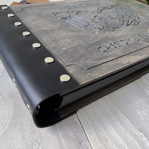 May include: A wooden photo album with a black leather spine and silver rivets. The cover is engraved with a design and the names "CASSIDY & SAMANTHA". The album is closed and appears to be new.