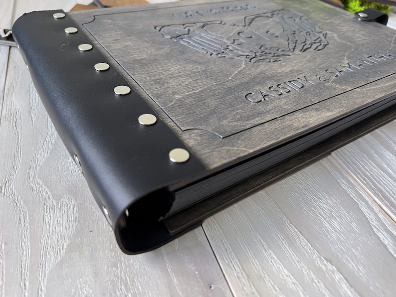 May include: A wooden photo album with a black leather spine and silver rivets. The cover is engraved with a design and the names "CASSIDY & SAMANTHA". The album is closed and appears to be new.