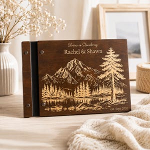 May include: A dark brown wooden photo album with a landscape engraving featuring a mountain range, a forest, and a lake. The cover is personalised with the text Stories in Strawberry, Rachel & Shawn, and est. Sept 2020, secured by metal rivets.