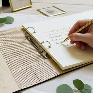 May include: A wooden scrapbook with a light-colored cover and a metal binder. A hand is writing with a gold pen on a cream-colored page. The scrapbook is open, revealing lined pages and decorative elements.