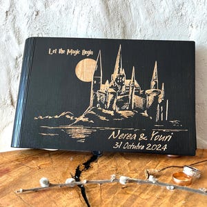 May include: Black wooden book with a laser-etched design of a castle, moon, and text "Let the Magic Begin." The names "Nerea & Youri" and the date "31 Octubre 2024" are also etched on the cover. The book is displayed on a wooden surface.