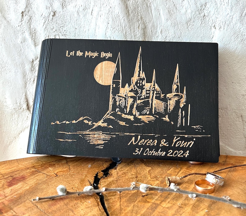 May include: Black wooden book with a laser-etched design of a castle, moon, and text "Let the Magic Begin." The names "Nerea & Youri" and the date "31 Octubre 2024" are also etched on the cover. The book is displayed on a wooden surface.