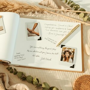 May include: An open wedding guest book with handwritten messages and polaroid photos. A gold pen rests on the page. Dried flowers and decorative elements surround the book, creating a warm, celebratory atmosphere.