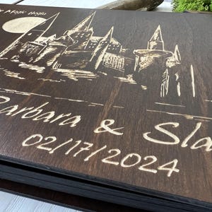 May include: A dark brown wooden guest book with a detailed engraving of a castle under a full moon. The names "Barbara & Slav" and the date "02/17/2024" are etched below the castle design. The text "Let the Magic Begin" is at the top.