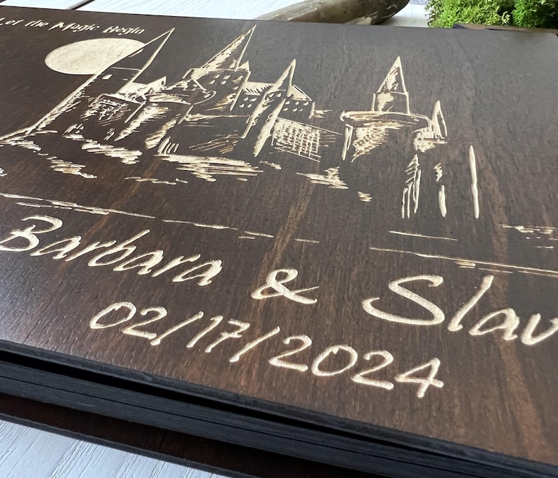 May include: A dark brown wooden guest book with a detailed engraving of a castle under a full moon. The names "Barbara & Slav" and the date "02/17/2024" are etched below the castle design. The text "Let the Magic Begin" is at the top.