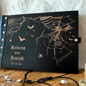 May include: Black wooden book with a leather spine and clasp, featuring a spiderweb and bat design. The names "Rebecca and Joseph" and the date "10/13/24" are engraved on the cover. A lit candle is in the background.