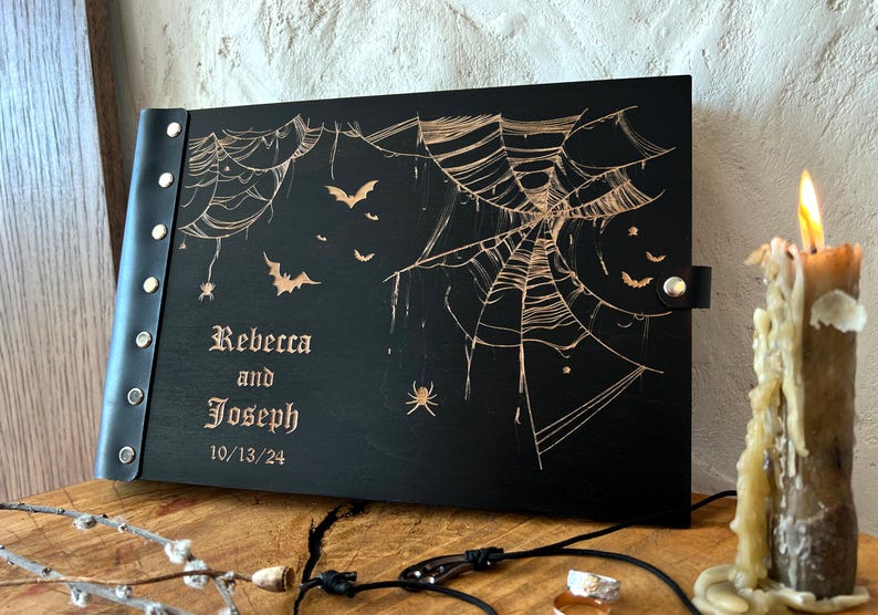 May include: Black wooden book with a leather spine and clasp, featuring a spiderweb and bat design. The names "Rebecca and Joseph" and the date "10/13/24" are engraved on the cover. A lit candle is in the background.