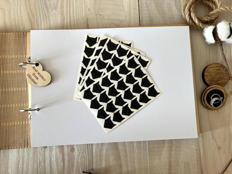 May include: An open scrapbook with white pages, a wooden cover, and a heart-shaped tag that reads "Best wishes from Ukraine!". Several black and white patterned stickers are on the page. Wedding rings are in a wooden box.