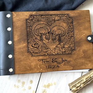 May include: A wooden photo album with a dark brown finish. The cover features an engraved design of two skulls surrounded by roses, with the names "Tom & Jen" and the date "1.5.24" below. The spine is black leather.