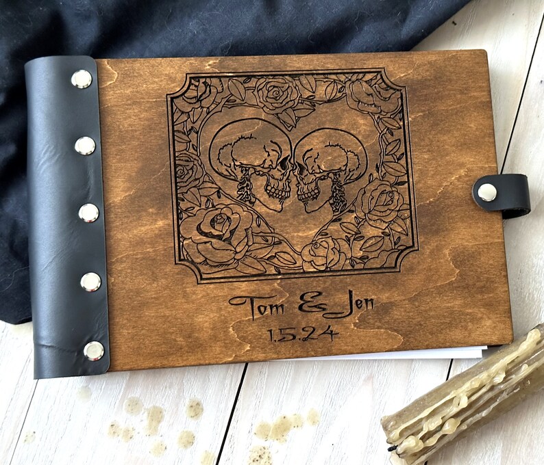 May include: A wooden photo album with a dark brown finish. The cover features an engraved design of two skulls surrounded by roses, with the names "Tom & Jen" and the date "1.5.24" below. The spine is black leather.