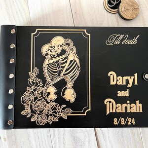May include: Black wooden photo album with a laser-cut design featuring embracing skeletons and roses. The album is personalized with the names "Daryl and Mariah" and the date "8/9/24". The words "Till Death" are also engraved on the cover.
