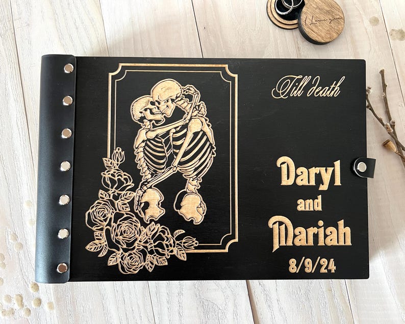 May include: Black wooden photo album with a laser-cut design featuring embracing skeletons and roses. The album is personalized with the names "Daryl and Mariah" and the date "8/9/24". The words "Till Death" are also engraved on the cover.