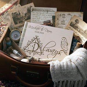 May include: A wooden scrapbook with the names "Michael & Dawn" and the date "03.05.2025" engraved on the cover, along with a Deathly Hallows symbol and an owl illustration. The scrapbook is in an open brown suitcase with vintage postcards and a compass.