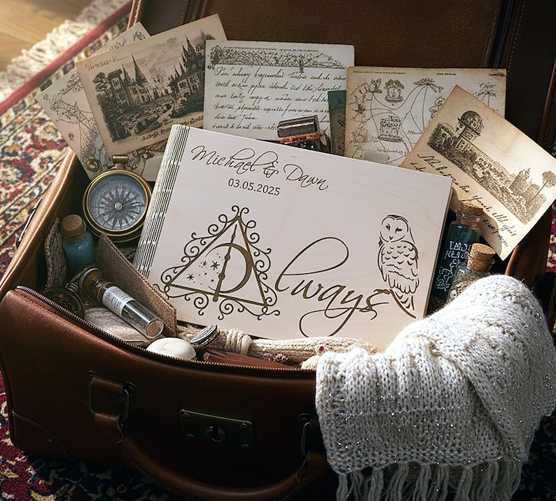 May include: A wooden scrapbook with the names "Michael & Dawn" and the date "03.05.2025" engraved on the cover, along with a Deathly Hallows symbol and an owl illustration. The scrapbook is in an open brown suitcase with vintage postcards and a compass.
