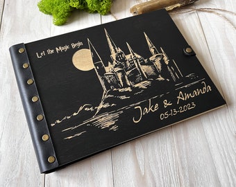 May include: A black wooden photo album with a gold engraved castle, a full moon, and the text "Let the Magic Begin" along with "Jake & Amanda" and "05-13-2023". The album features a black leather spine secured with six brass-colored metal rivets.
