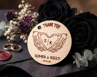 Può includere: Un tag rotondo in legno con le parole "THANK YOU" in alto. Il tag presenta un cuore fatto di mani di scheletro, con le iniziali "S | K" all'interno. I nomi "SUMER & KELLY" e la data "10/31/26" sono in basso.