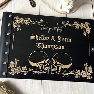 May include: Black wooden wedding guest book with a leather spine and silver rivets. The cover features a floral design and two skulls, with the text "I love you to death" and the names "Shelby & Jenn Thompson" engraved in gold.