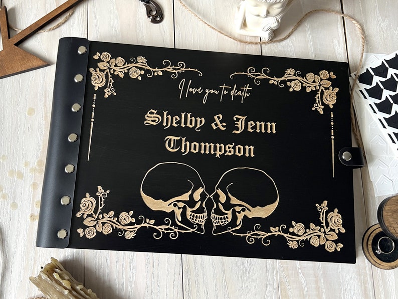 May include: Black wooden wedding guest book with a leather spine and silver rivets. The cover features a floral design and two skulls, with the text "I love you to death" and the names "Shelby & Jenn Thompson" engraved in gold.