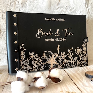 Może przedstawiać: Czarna skórzana księga gości z pozłacanymi napisami. Księga zawiera napis "Our Wedding" oraz "Barb & Tim" z datą "October 5, 2024". Okładka zdobiona jest wzorem kwiatowym z kwiatem hibiskusa.