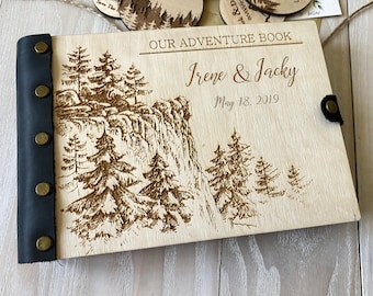 Peut inclure: Un album photo en bois avec une reliure en cuir noir fixée par cinq rivets en laiton. La couverture présente une gravure laser d'un paysage de falaise avec des pins, le texte "OUR ADVENTURE BOOK", les noms "Irene & Jacky" et "May 18, 2019".