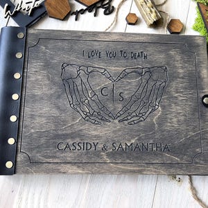 May include: A gray wooden photo album with a black leather spine and clasp. The cover is engraved with skeleton hands forming a heart, the text "I LOVE YOU TO DEATH", and the names "CASSIDY & SAMANTHA".