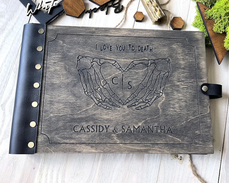 May include: A gray wooden photo album with a black leather spine and clasp. The cover is engraved with skeleton hands forming a heart, the text "I LOVE YOU TO DEATH", and the names "CASSIDY & SAMANTHA".