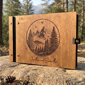 May include: A rectangular wooden photo album with a dark brown finish. The front cover features a circular laser-engraved mountain and pine tree landscape. The text "October 31, 2025" and "Jonathan and Samantha" are engraved on the cover.