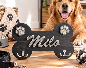 Könnte beinhalten: Ein Wandhaken aus dunklem Holz in Knochenform mit dem Namen Milo in erhabener silberner Schrift. Das Stück verfügt über zwei silberne Pfotenabdrücke oben und zwei silberne Metallhaken unten zum Aufhängen von Gegenständen.