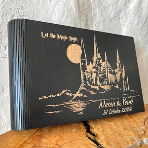 May include: A black wooden box with a laser-etched design featuring a castle, moon, and text that reads "Let the Magic Begin." The box also includes the names "Nerea & Youri" and the date "31 Octubre 2024."