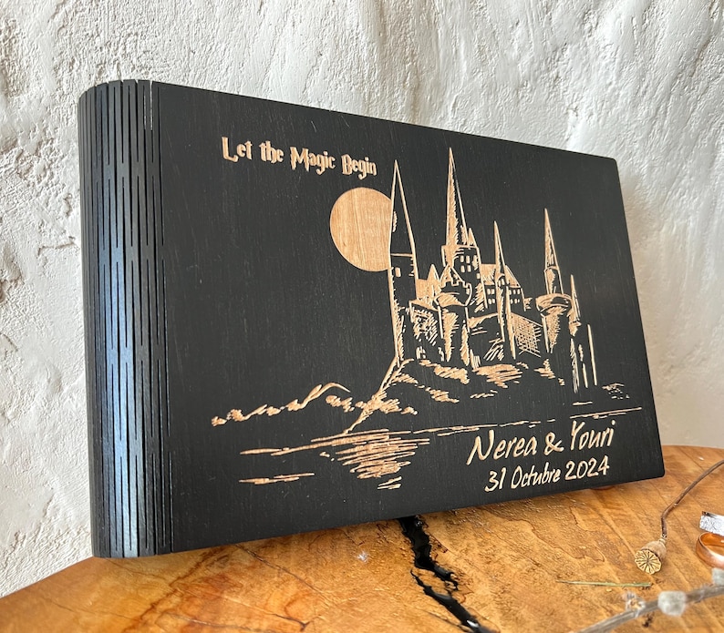 May include: A black wooden box with a laser-etched design featuring a castle, moon, and text that reads "Let the Magic Begin." The box also includes the names "Nerea & Youri" and the date "31 Octubre 2024."