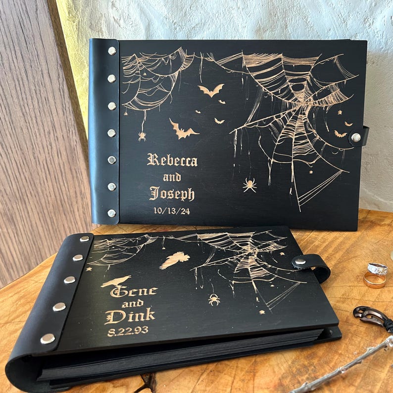 May include: Two black leather-bound photo albums with engraved spiderwebs, bats, and spiders. One album reads "Rebecca and Joseph 10/13/24", the other reads "Gene and Dink 8.22.93". Both have silver rivets and a leather strap closure.