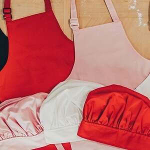 May include: Four chef hats in different colours: black, red, white, and pink. The hats are made of a soft, fabric material. There are also three aprons in the background: red, pink, and white.