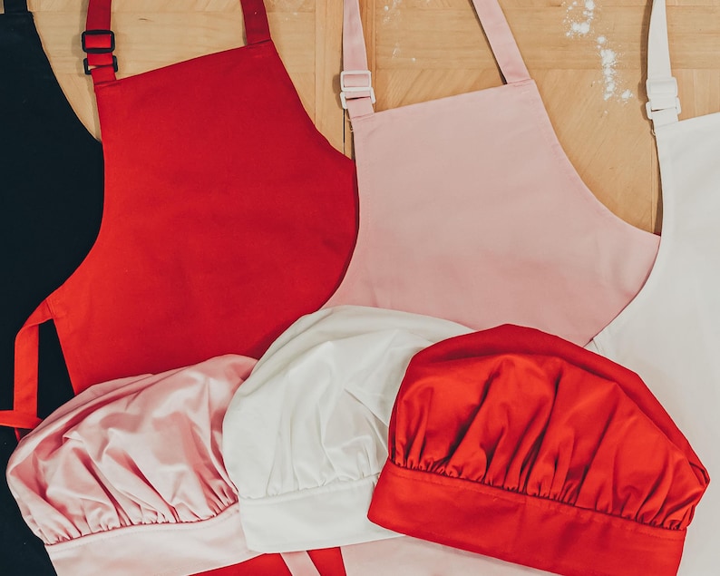 May include: Four chef hats in different colours: black, red, white, and pink. The hats are made of a soft, fabric material. There are also three aprons in the background: red, pink, and white.