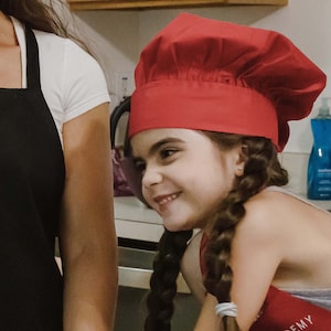 May include: A young girl wearing a red chef's hat and a grey shirt with the word "EMY" on it. She is smiling and looking to the side.