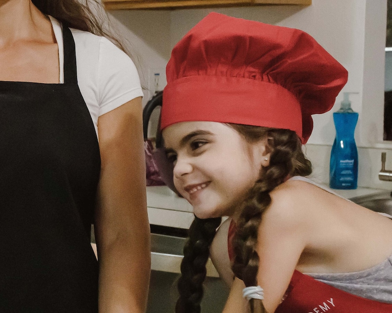 May include: A young girl wearing a red chef's hat and a grey shirt with the word "EMY" on it. She is smiling and looking to the side.