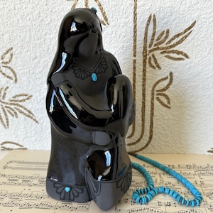 May include: A black ceramic figurine depicting a woman and child, embellished with turquoise details. The glossy sculpture has intricate patterns and is accompanied by a turquoise beaded necklace. The figurine rests on sheet music.