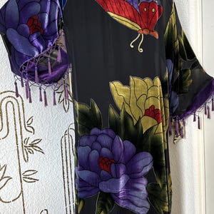 May include: Black kimono-style robe with a floral and butterfly design. The robe features large purple and yellow flowers, a butterfly with red and yellow wings, and purple velvet trim with tassels. The sleeves and collar are trimmed with purple velvet.