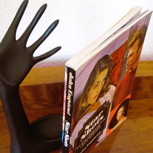 May include: A black hand-shaped bookend supports a book titled "Star Shots" by John Engstead. The book cover features images of people in a vintage style. The bookend is on a wooden surface.