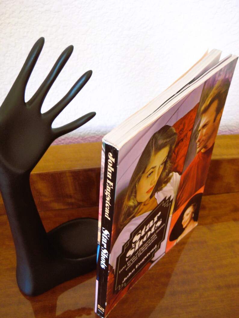 May include: A black hand-shaped bookend supports a book titled "Star Shots" by John Engstead. The book cover features images of people in a vintage style. The bookend is on a wooden surface.