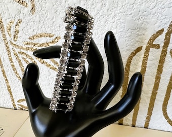 May include: A vintage bracelet featuring alternating rows of black rectangular stones and clear, sparkling rhinestones. The bracelet is displayed on a black hand-shaped display, showcasing its elegant design. The background has a textured white surface with gold accents.