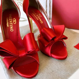 Puede incluir: Sandalias de tac&oacute;n alto de sat&eacute;n rojo con lazos. Las sandalias tienen tacones dorados y la marca "Valentino Garavani" es visible en la plantilla. Los zapatos se exhiben sobre una superficie de color neutro.