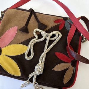 Puede incluir: Bolso de hombro de ante con diseño floral en tonos marrón, rojo, rosa y amarillo. El bolso tiene una correa roja y un collar de cuentas. El bolso mide aproximadamente 30 cm de ancho y 25 cm de alto.