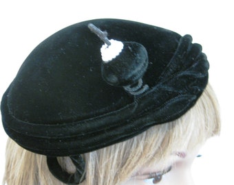 May include: A black velvet hat with a black and white trim. The hat has a small, round, black velvet ball on top.